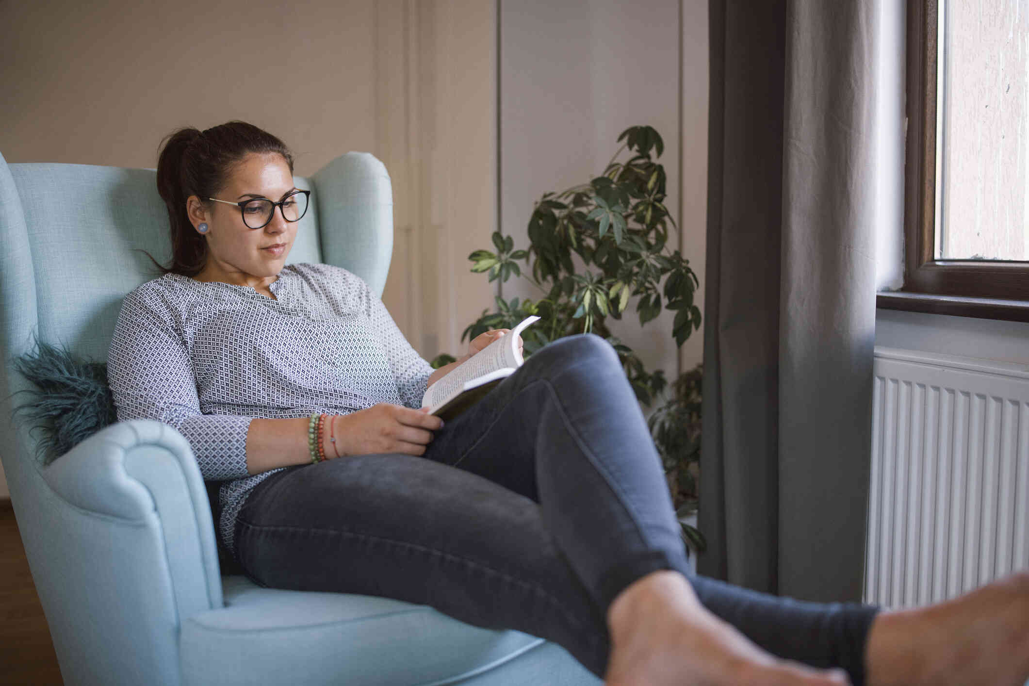A woman with glasses sits in a blue armchair in her home and reads  from the book in her lap.